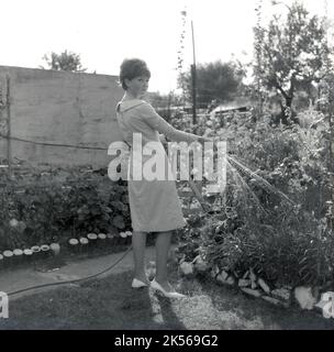 1964, historique, l'été et à l'extérieur dans un jardin, une jeune femme dans une robe arroser ses plantes, en utilisant une pipe à hope avec un spray, Angleterre, Royaume-Uni. Banque D'Images