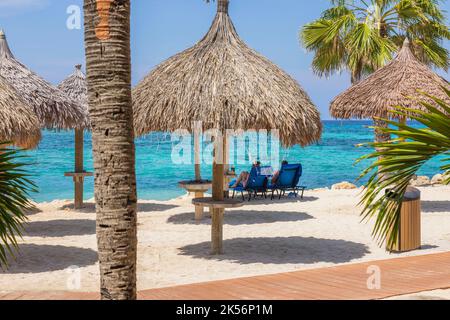 Vue magnifique sur un jeune couple sous des parasols sur une plage de sable blanc de l'île d'Aruba sur fond d'eau turquoise de l'océan Atlantique. Banque D'Images