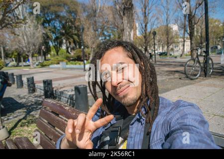 le jeune vénézuélien latin avec des dreadlocks est assis dans une place à l'extérieur, il prend un selfie souriant et faisant un signe v avec sa main, signe de paix Banque D'Images