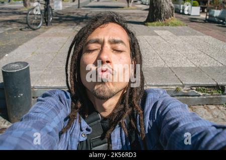 jeune touriste vénézuélien latin avec des dreadlocks et des vêtements décontractés, prend un selfie et envoie un message photo avec un baiser à sa petite amie. Banque D'Images