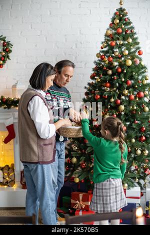fille décorant l'arbre de noël avec des grands-parents interraciaux dans la salle de séjour, image de stock Banque D'Images