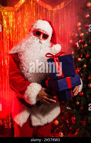Le Père noël en lunettes de soleil tenant des cadeaux près de l'arbre de noël et scintillant, image de stock Banque D'Images