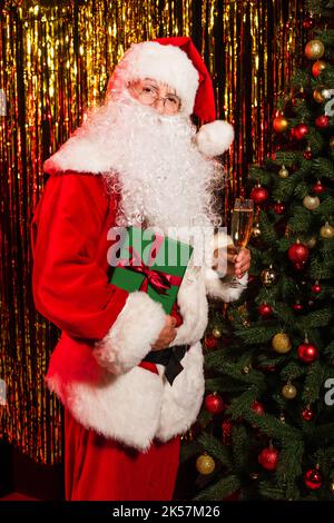 Le Père noël en lunettes tenant le champagne et le présent près de l'arbre de noël et de la guirlande, image de stock Banque D'Images