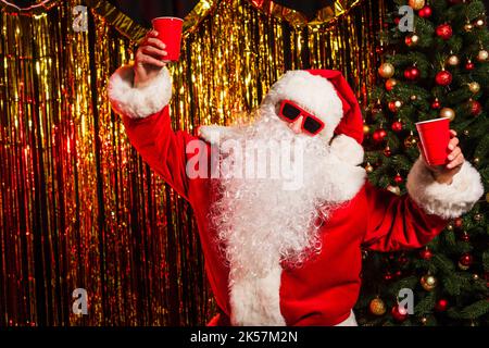 Le Père noël en lunettes de soleil tenant des tasses jetables près de l'arbre de noël et de la guirlande, image de stock Banque D'Images