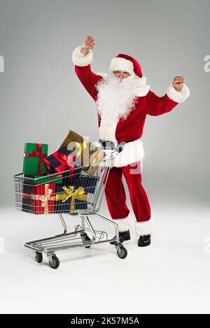 Pleine longueur du père noël excités debout près du panier avec des cadeaux sur fond gris, image de stock Banque D'Images