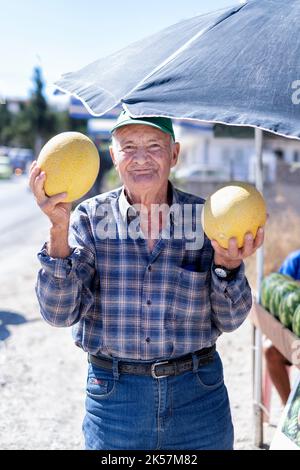 Un homme âgé tenant deux grands melons jaunes de miel Cucumis melo. L'homme vend les melons d'un étal au bord de la route à Rhodes Banque D'Images