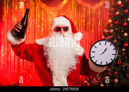 Le Père noël en lunettes de soleil tenant une bouteille de champagne et une horloge près de l'arbre de noël, image de stock Banque D'Images