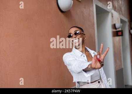 Joyeuse et élégante femme afro-américaine montrant le signe de la paix sur la rue urbaine, image de stock Banque D'Images