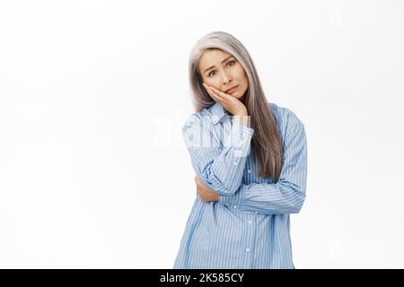 Image d'une femme asiatique âgée triste et sombre, qui regarde troublée et inquiète la caméra, se tenant contrariée par un arrière-plan blanc Banque D'Images