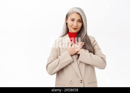 Portrait d'une femme d'affaires asiatique heureuse, femme d'affaires senior en costume, en regardant reconnaissant, en tenant les mains sur le coeur, geste de remerciement, debout sur blanc Banque D'Images
