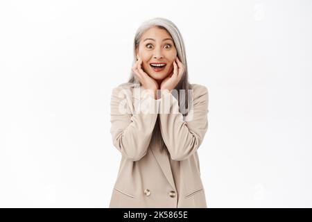 Portrait d'une femme asiatique heureuse semble surpris, souriant enthousiaste et regardant l'appareil photo, portant un costume élégant, debout sur fond blanc Banque D'Images