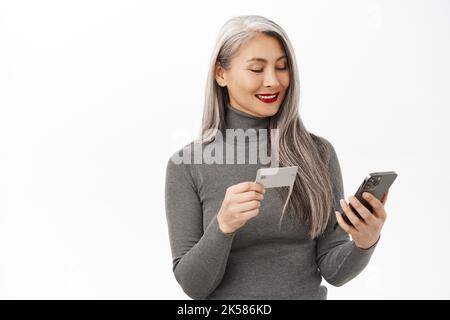 Belle femme asiatique senior utilisant le téléphone mobile, tenant une carte de crédit, commander ou faire des achats en ligne, debout heureux sur fond blanc Banque D'Images