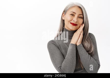 Belle femme japonaise d'âge moyen, portant un maquillage rouge à lèvres, souriant heureux, tenant les mains près d'un visage sain et frais, debout sur blanc Banque D'Images