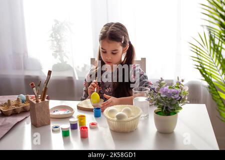 Une adorable petite fille s'assoit à une table blanche près de la fenêtre, peint des œufs Banque D'Images