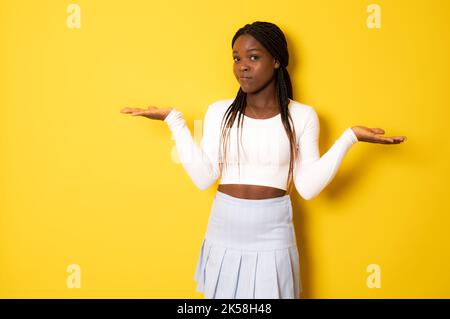 Jeune afro-américaine isolée sur fond jaune doutant et haussant les épaules dans le geste de questionnement. Banque D'Images