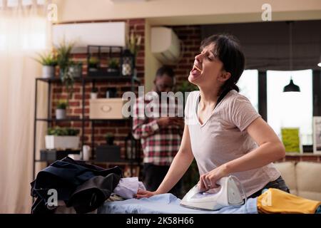 Bonne femme souriante chantant et repassant des vêtements à bord, mari au téléphone en arrière-plan. Culturel couple divers à la maison, femme au foyer gaie faisant des tâches quotidiennes avec des vibes positives et des émotions Banque D'Images