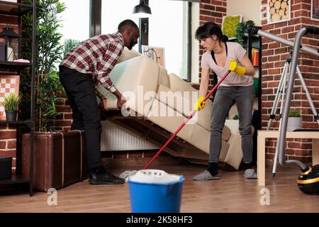 Culture diverse couple de nettoyage de printemps l'appartement, le mari monte le canapé et la femme au foyer est humide de laver le sol en dessous. Nettoyage de la maison et travaux ménagers de jour, lavage et passer l'aspirateur avec du détergent. Banque D'Images