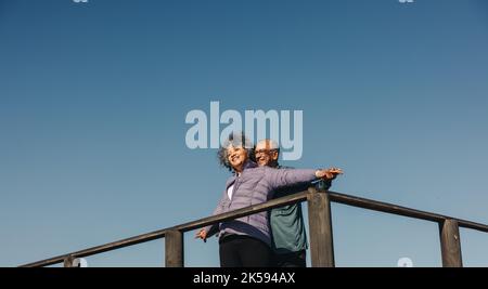 Joyeux couple senior souriant et étalant leurs bras tout en se tenant au sommet d'un pont de pied en bois. Couple romantique de personnes âgées s'amuser sur un rel Banque D'Images