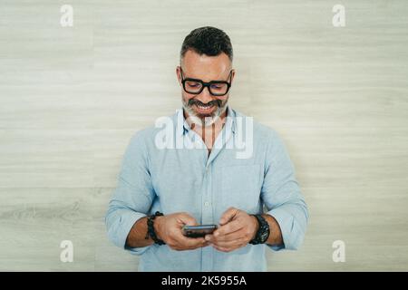 Homme d'affaires mûr souriant gaiement tout en lisant un message texte sur son smartphone. Homme d'affaires réussi à communiquer avec ses associés pendant que W Banque D'Images