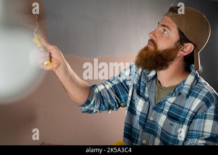 Peintre barbu avec un rouleau de peinture près du mur, peint le mur haut. Réparation, bâtiment et maison Banque D'Images