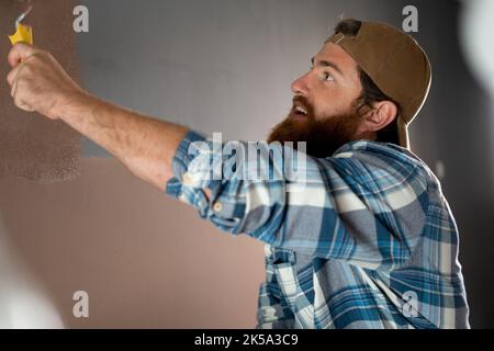 Peintre de maison barbu tenant un rouleau de peinture et peint un mur dans une maison. Réparation, bâtiment et maison Banque D'Images