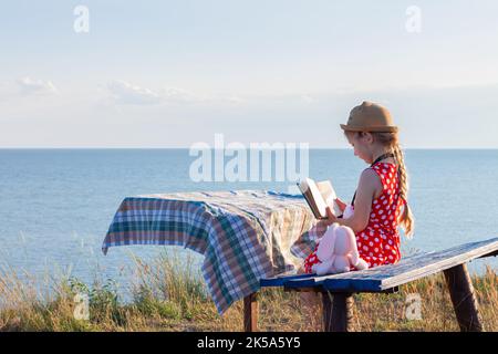 Enfant fille en chapeau et robe à pois assis sur un banc vintage lisant un livre. Enfant regardant la Sainte Bible dans les mains et priant sur la mer lanscape backgrou Banque D'Images