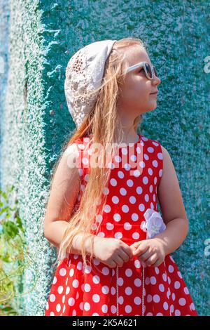 Belle fille heureuse dans une robe à pois rouges posant et souriant sur fond de mur en béton turquoise.mignon enfant joyeux avec de longs cheveux blonds chantés Banque D'Images