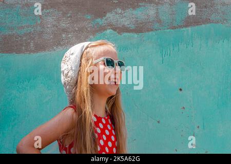 Belle fille heureuse dans une robe à pois rouges posant et souriant sur fond de mur en béton turquoise.mignon enfant joyeux avec de longs cheveux blonds chantés Banque D'Images