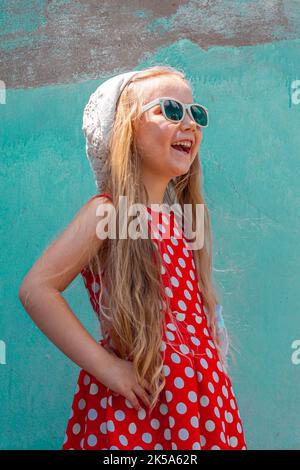 Belle fille heureuse dans une robe à pois rouges posant et souriant sur fond de mur en béton turquoise.mignon enfant joyeux avec de longs cheveux blonds chantés Banque D'Images