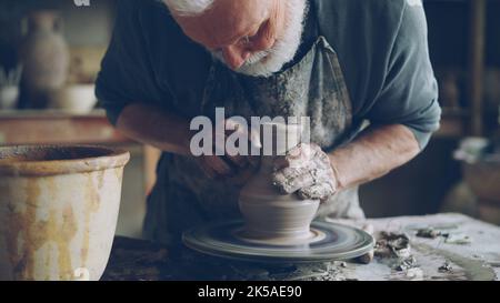 Gros plan d'un pot en céramique semi-fini tournant sur la roue des potiers et les mains mâles moulant l'argile avec des outils professionnels. Création de la faïence et de la poterie traditionnelle. Banque D'Images