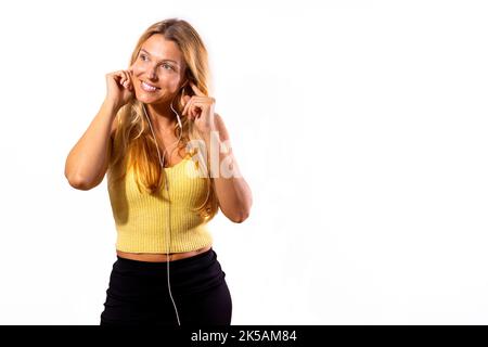 Une jeune fille hispanique blonde écoute de la musique avec un casque. Cheveux blonds amples et vêtements décontractés avec fond blanc. Banque D'Images