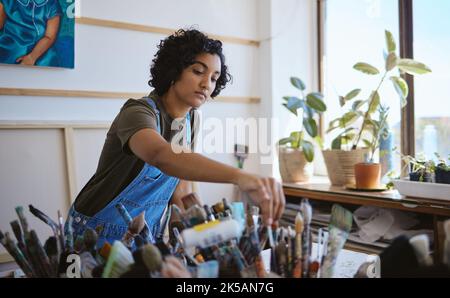 Art, créatif et peintre avec la décision de pinceau tout en travaillant sur une peinture dans un studio pour le travail. Femme indienne, artiste ou designer choisissant un outil Banque D'Images