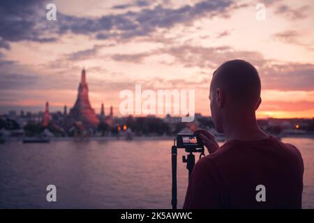 Homme photographiant le paysage urbain au coucher du soleil. Tourisme avec appareil photo sur trépied contre le temple de Wat Arun à Bangkok, Thaïlande. Banque D'Images