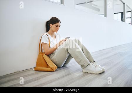 Triste femme universitaire, étudiante seule sur le sol dans le couloir après la classe ou la conférence et la navigation sur les médias sociaux sur smartphone. Dépression universitaire Banque D'Images