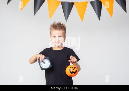 Joyeux Halloween dans un T-shirt noir tenant un seau en forme de citrouille avec des friandises et un réveil noir, sur un fond blanc avec une guirlande. Hall Banque D'Images