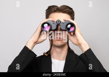 Portrait d'un jeune homme élégant portant un costume noir regardant à travers des jumelles. Studio tourné sur fond gris Banque D'Images