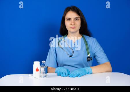 Un médecin en uniforme bleu avec un stéthoscope et des gants est assis à une table avec des médicaments. Banque D'Images