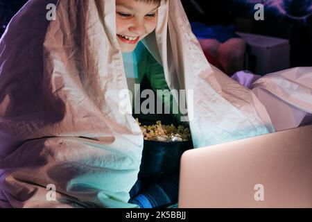 Un enfant heureux, un fils riant, mange du pop-corn à télécommande en regardant une comédie télé amusante, assis sur un canapé et en regardant des vidéos sur un ordinateur portable le soir à la maison. Banque D'Images