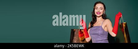 bonne jeune femme en gants rouges et robe violette tenant des sacs de shopping sur le vert, bannière, image de stock Banque D'Images