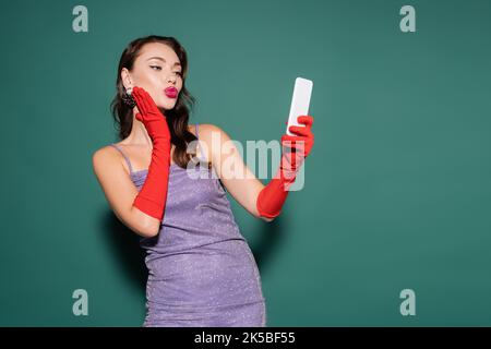 jeune femme en robe pourpre et gants poudant les lèvres tout en prenant selfie sur smartphone sur vert, image de stock Banque D'Images