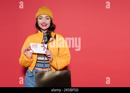 jeune femme gaie en bonnet beanie orange tenant une carte-cadeau et un sac à provisions sur rouge, image de stock Banque D'Images