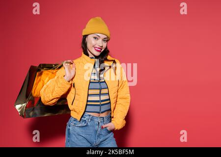 jeune femme gaie en bonnet beanie orange et veste bouffante tenant le sac de shopping sur rouge, image de stock Banque D'Images