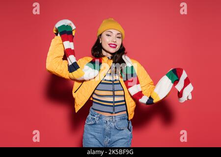 jeune femme gaie en bonnet beanie et blouson orange tenant une écharpe striée sur rouge, image de stock Banque D'Images