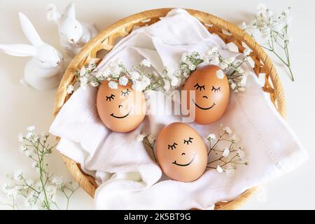 Trois oeufs de Pâques heureux avec des visages mignons dans des couronnes de couronne florales sur fond blanc dans le nid et deux lapins blancs. Oeufs de Pâques avec fleurs et somnolent Banque D'Images