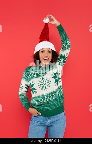 joyeuse femme afro-américaine tenant le pompon sur le chapeau de santa tout en se tenant avec la main dans la poche isolée sur rouge, image de stock Banque D'Images
