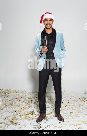 joyeux homme américain africain en blazer de soie et chapeau de père noël debout avec coupe de champagne sur confetti sur fond gris, image de stock Banque D'Images