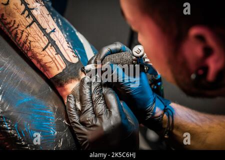 Portrait de l'homme tatouage maître avec des dreadlocks montrant le processus de création tatouage sur le corps femelle sous la lumière de lampe. Artiste professionnel travaillant dans un salon Banque D'Images