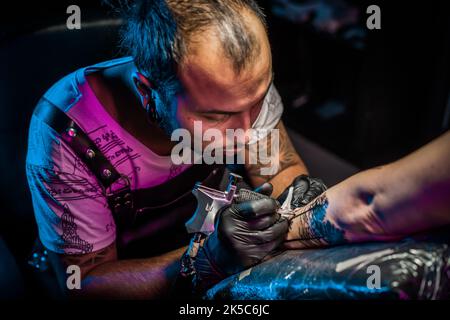 Portrait de l'homme tatouage maître avec des dreadlocks montrant le processus de création tatouage sur le corps femelle sous la lumière de lampe. Artiste professionnel travaillant dans un salon Banque D'Images