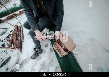 Un homme verse une tasse de café d'un thermos sur un banc vert à l'extérieur et à côté d'une boîte de dessert français traditionnel eclair. Pique-nique en hiver Banque D'Images