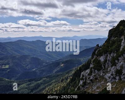 Vue spectaculaire depuis la chaîne de montagnes de Raxalpe en Autriche, avec des falaises rocheuses abruptes, des vallées boisées, des montagnes bleues en couches et un ciel alpin nuageux. Banque D'Images